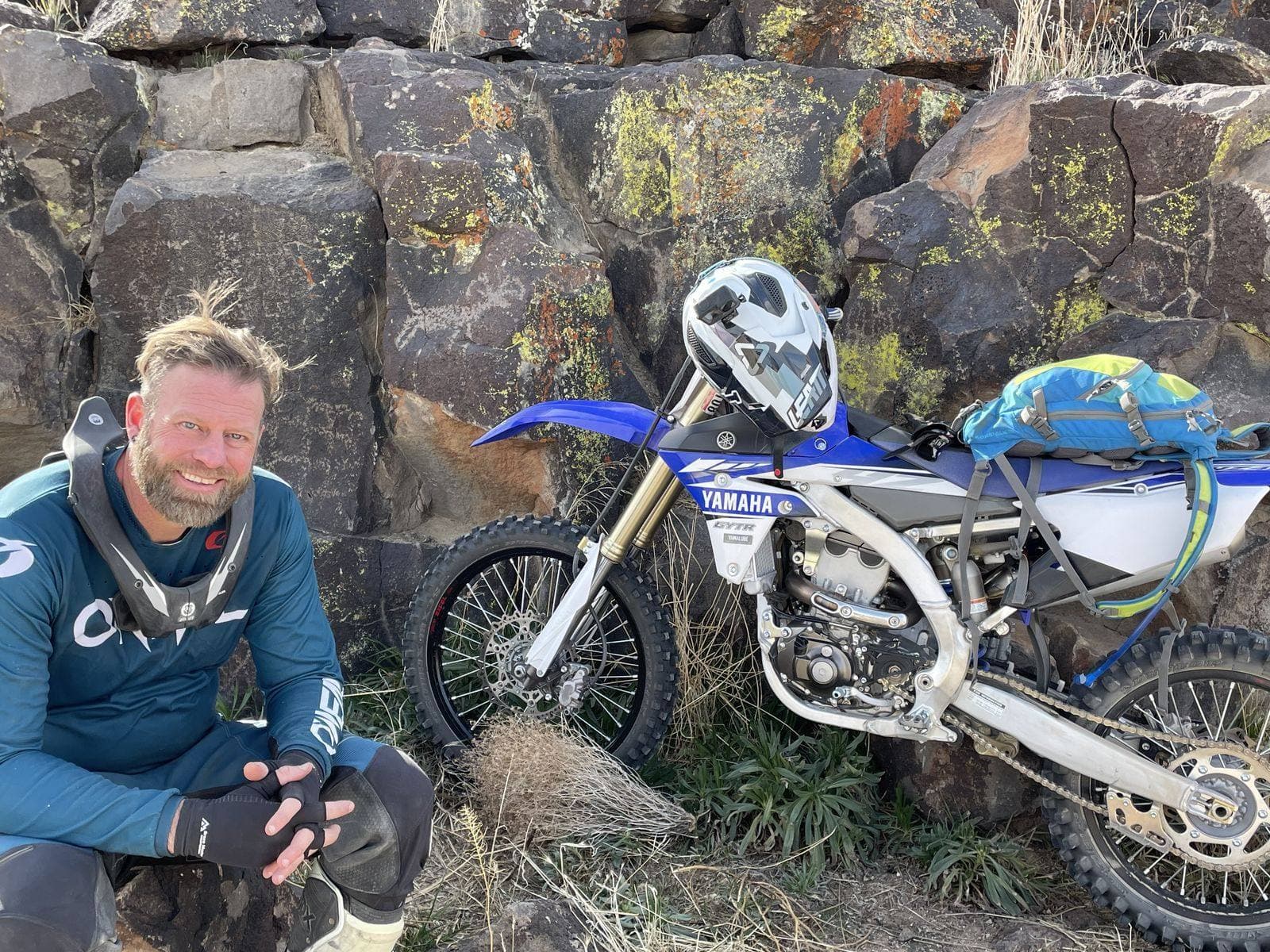 A rider sitting on the rocks beside his dirt bike, gloves off, at a stop on the ride