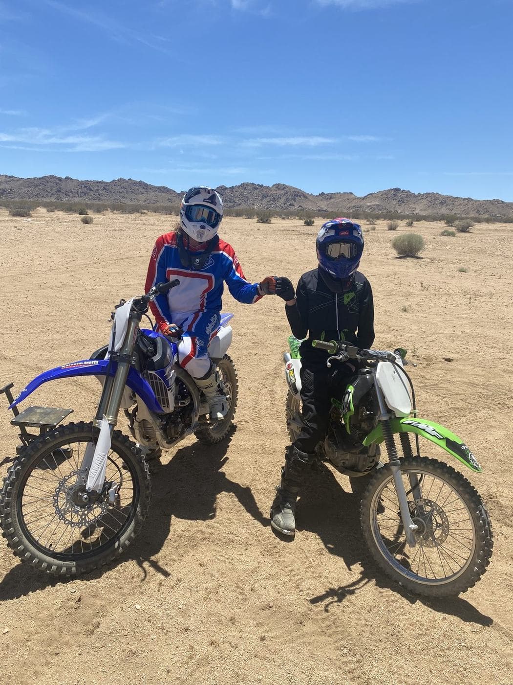 Two helmeted riders on dirt bikes bumping fists on a desert flat
